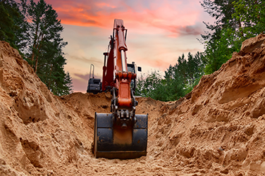 Excavator equipment digging into dirt with sunset and trees in the background
