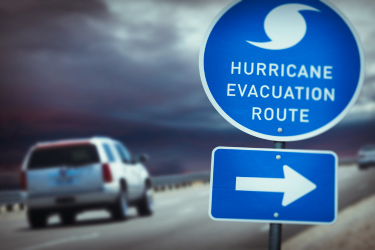  A hurricane evacuation route road sign in the foreground with cars on a road against a dark sky in the background.